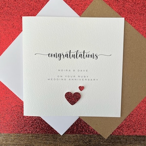 May include: A square white greeting card with black text that reads congratulations MOIRA & DAVE ON YOUR RUBY WEDDING ANNIVERSARY. The card features two small red glittery heart embellishments positioned below the printed text.