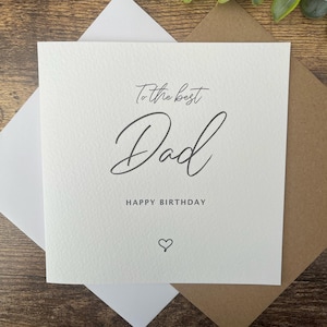 May include: A square white greeting card with black text that reads To the best Dad, HAPPY BIRTHDAY, and a small heart symbol at the bottom. The card is positioned on top of a white envelope and a brown envelope, with a green leaf sprig visible.