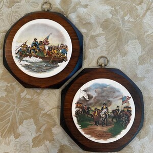 May include: Two octagonal wooden wall plaques featuring circular ceramic inserts. One depicts Washington Crossing the Delaware by Emanuel Leutze, and the other shows the Surrender of Cornwallis by John Trumbull, both with metal hanging rings.