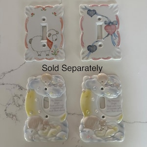 May include: Four rectangular ceramic light switch plates with decorative edges. Two plates feature painted designs of a sheep and hearts with ribbons. Two plates feature embossed figures of children on clouds with the text My Precious Angel.