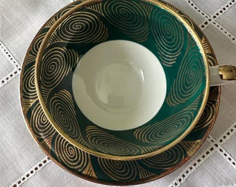 May include: An overhead shot of a teacup and saucer set. The teacup and saucer are dark green with gold spiral designs and gold trim. The inside of the teacup is white. The set is on a white, patterned tablecloth.