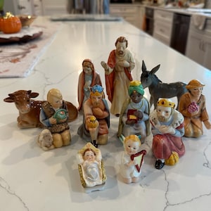 May include: A ceramic nativity set featuring eleven hand-painted figurines including a central infant in a manger, an angel with a harp, three kings, a shepherd with a lamb, a brown cow, a donkey, and two other figures in traditional robes.