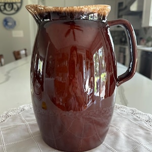 May include: A large, glossy brown pitcher with a handle and a spout. The pitcher has a decorative pattern near the base and a lighter brown rim. The pitcher is sitting on a white lace tablecloth.