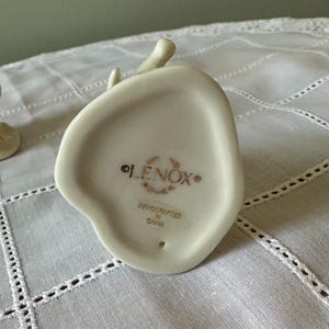 May include: The underside of an off-white porcelain figurine featuring the gold-colored text LENOX, surrounded by a decorative wreath emblem, with the words HANDCRAFTED IN CHINA printed below. A small circular hole is visible at the bottom.