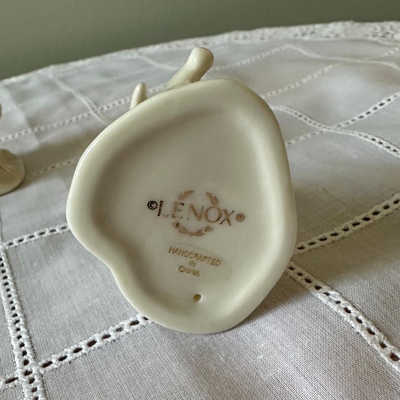 May include: The underside of an off-white porcelain figurine featuring the gold-colored text LENOX, surrounded by a decorative wreath emblem, with the words HANDCRAFTED IN CHINA printed below. A small circular hole is visible at the bottom.