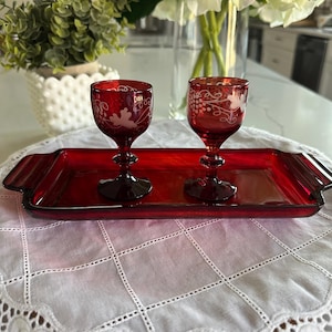 May include: Two ruby red wine glasses with etched grape designs sit on a matching rectangular tray. The tray has handles on each end. The glasses and tray are on a white tablecloth.