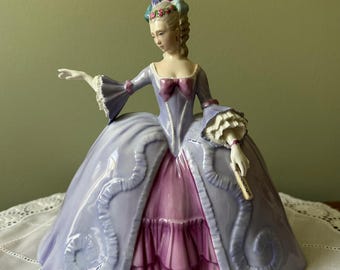 May include: A porcelain figurine of a woman in a period gown. The dress is a light purple with pink accents and a ruffled skirt. The woman has a powdered wig with colorful accents and is holding a fan. The figurine is a decorative item.