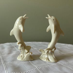 May include: Two ivory-colored ceramic dolphin figurines. Each dolphin is leaping from a wave base, with gold trim accents. The dolphins have open mouths and are posed as if they are jumping out of the water. Decorative home decor.