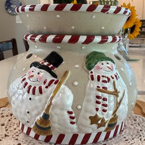May include: A ceramic wax warmer featuring a cream-colored base with white polka dots and red and white striped trim. The exterior is decorated with two raised snowman figures, one wearing a black top hat and the other a green hat, both holding brooms.