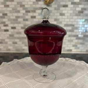 May include: A ruby red glass apothecary jar with a clear glass lid and stem. The jar has a rounded body and a clear, textured knob on top. The stem is clear glass, and the jar sits on a round base. The jar is likely used for storing items.