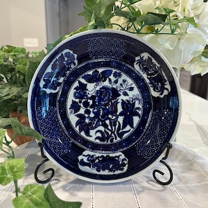 May include: A blue and white decorative plate with a floral design. The plate features a central floral motif surrounded by a dotted border and floral accents. The plate is displayed on a black metal stand.