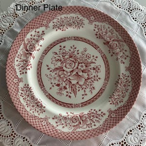 May include: A white dinner plate with a pink floral design. The plate features a central bouquet of roses and other flowers, surrounded by a decorative border and a lattice pattern on the rim. The text "Dinner Plate" is at the top.