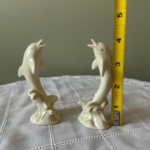 May include: Two ivory-colored ceramic dolphin figurines, each approximately 4 inches tall, leaping from stylized waves. The dolphins have open mouths and are accented with gold trim, creating a decorative nautical theme.