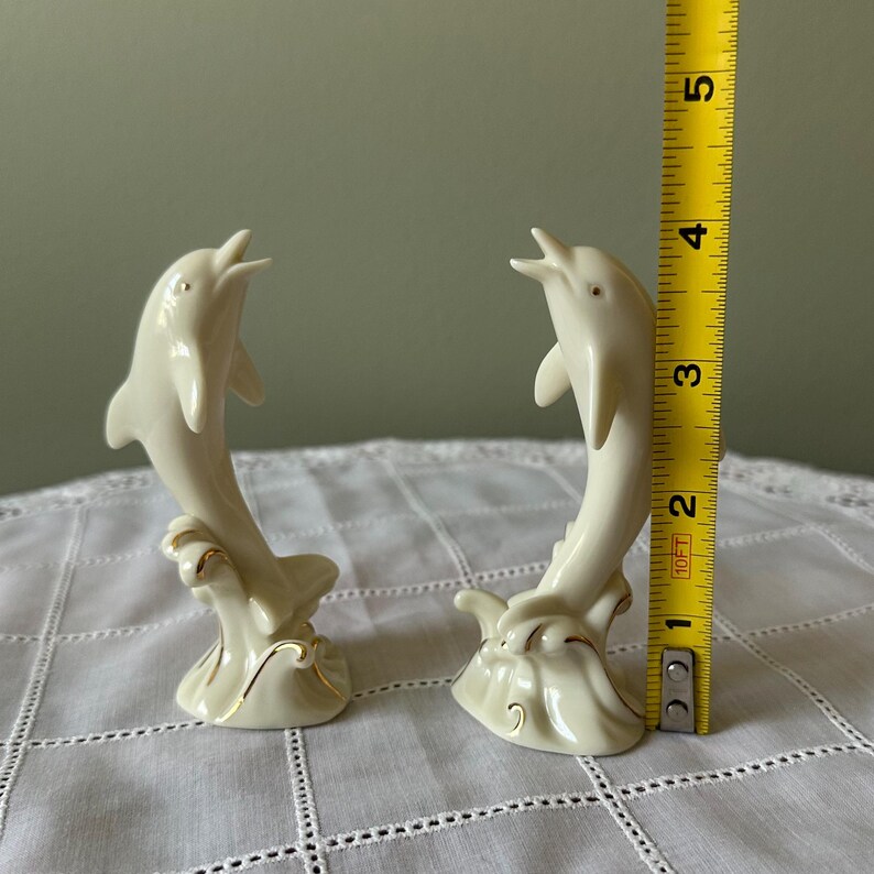 May include: Two ivory-colored ceramic dolphin figurines, each approximately 4 inches tall, leaping from stylized waves. The dolphins have open mouths and are accented with gold trim, creating a decorative nautical theme.