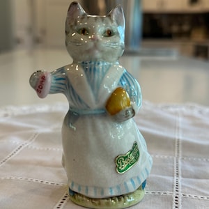 May include: A ceramic figurine of a cat wearing a blue and white striped dress and apron, holding a yellow object and a pink soap. The figurine has a green and gold "Beswick England" sticker.