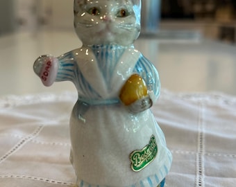 May include: A ceramic figurine of a cat wearing a blue and white striped dress and apron, holding a yellow object and a pink soap. The figurine has a green and gold "Beswick England" sticker.