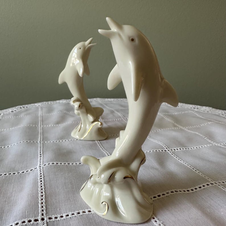 May include: Two ivory-colored ceramic dolphin figurines. The dolphins are leaping out of stylized waves, with gold accents on the base. One dolphin is larger than the other. The figurines are displayed on a white, textured tablecloth.