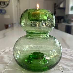 May include: A green glass candle holder with a double-bulb, rounded shape and a textured, bubbly surface. The top section features a recessed circular opening holding a small, lit tea light candle, while the base is wide and rounded.