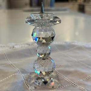 May include: A clear crystal candle holder featuring a square base and a square top platform with a central metal spike. The stem consists of three stacked, faceted spherical crystal beads of varying sizes, creating a reflective, geometric design.