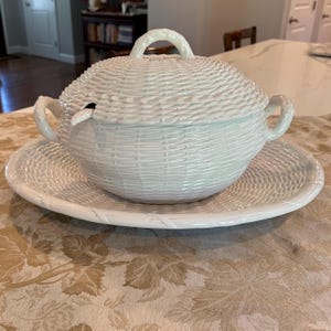 May include: A white ceramic tureen with a matching lid and oval platter, featuring a textured basket-weave pattern throughout. The tureen has two side handles and a central lid handle, with a small opening for a ladle on the side of the bowl.