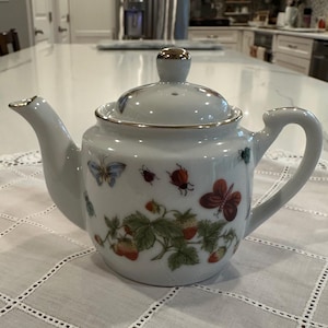 May include: A white porcelain teapot featuring a delicate botanical pattern of red strawberries, green leaves, and various colorful butterflies and ladybugs. The teapot includes a matching lid with a gold-trimmed knob, a curved spout, and a handle.
