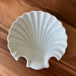 May include: A white, shell-shaped dish with a scalloped edge. The dish has a glossy finish and is made of ceramic. The dish is designed to resemble a seashell and is likely used for serving small items or as a decorative piece.