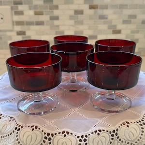 May include: Six ruby red glass dessert bowls with clear stems and bases are arranged on a white lace tablecloth. The bowls have a wide, shallow design. The background features a neutral-toned tile backsplash.