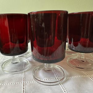 May include: A collection of ruby red stemmed glasses. The glasses have a cylindrical bowl and a clear, circular base. The stem is short and clear. The glasses are arranged on a white tablecloth.