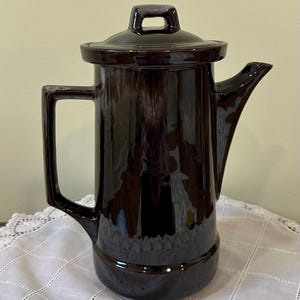 May include: A tall, cylindrical ceramic coffee pot with a glossy dark brown glaze. It features a sturdy angular handle, a curved pouring spout, and a matching lid with a small knob handle. The base has a subtle embossed grid pattern.