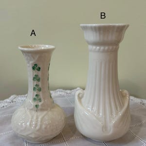 May include: Two off-white ceramic vases labeled A and B. Vase A is smaller with a bulbous base and a vertical row of green shamrock patterns. Vase B is taller with a fluted neck, a wide rim, and decorative scroll accents at the base.