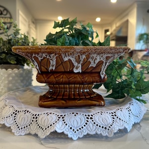 May include: A brown ceramic planter with a rectangular top and a pedestal base. The planter has a textured surface with white accents along the rim. It sits on a white lace doily, with green plants in the background.