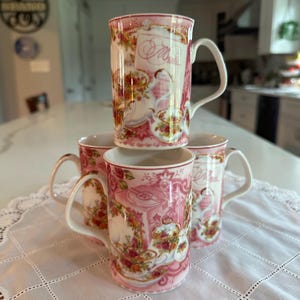 May include: Four stacked pink ceramic mugs with white handles. Each mug features a floral and tea-themed design with the word "Menu" visible on one mug. The mugs are arranged on a white lace tablecloth.
