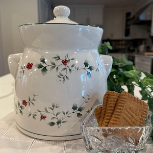 May include: A white ceramic cookie jar with a lid, decorated with green holly leaves and red berries. The jar has two handles and a green trim. A small glass dish holds several brown cookies.