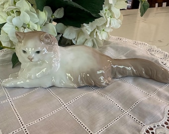 May include: A porcelain figurine of a long-haired cat in a reclining position. The cat features a glossy finish with a cream-colored body and light brown shading on the ears, face, and long, flowing tail, capturing a realistic feline form.