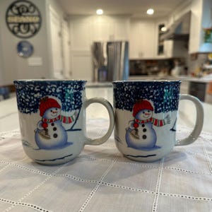 May include: Two ceramic mugs featuring a hand-painted snowman wearing a red hat and a red and green striped scarf. The upper portion of each mug has a dark blue speckled pattern, while the lower section is white with a snowman illustration.