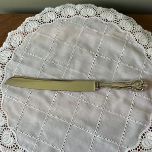 May include: A silver-colored cake knife with a decorative handle rests on a white, square-patterned doily with a scalloped lace edge. The knife blade is smooth and the handle has an ornate floral design.