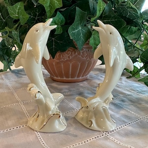 May include: A pair of cream-colored ceramic dolphin figurines, each depicted leaping from a stylized wave base. The dolphins feature smooth, glossy surfaces with delicate gold-painted trim along the edges of the waves, creating a refined and elegant appearance.
