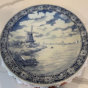 May include: A round ceramic decorative plate featuring a blue and white transferware scene of a Dutch windmill by a river. The border displays a detailed floral pattern. The plate includes the text 'Delft' and 'Made in Chantal' on the surface.
