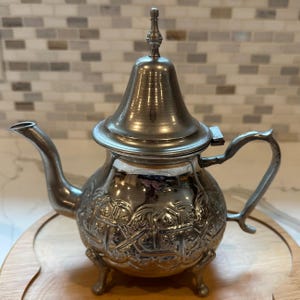 May include: A silver-toned metal teapot featuring a rounded body with intricate embossed geometric patterns, a curved spout, a hinged lid with a pointed finial, and three ornate legs. The polished surface reflects light across its detailed exterior.