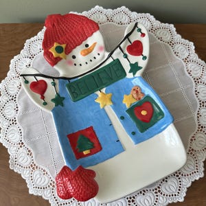 May include: A snowman-shaped ceramic serving platter with a red knit hat and mittens. The snowman has a blue vest with the word "BELIEVE" and decorative hearts, stars, and a gingerbread man. A festive holiday decoration.