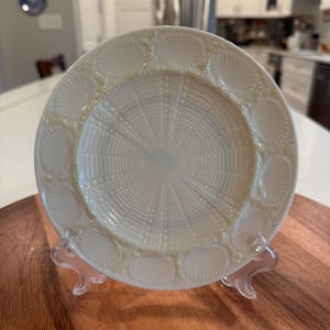 May include: A round, off-white ceramic plate featuring a central radial pattern of raised lines and dots. The rim is decorated with a repeating series of embossed shell motifs, each separated by small, pearl-like beaded accents in a glossy finish.