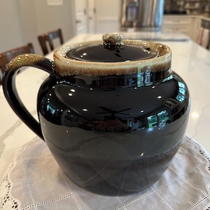 May include: A black ceramic bean pot with a lid and handle. The pot has a glossy finish with a brown and tan rim and handle. The pot is sitting on a white cloth.