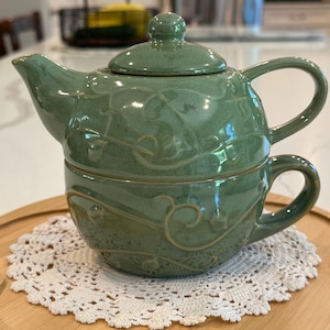 May include: A sage green ceramic tea set for one, featuring a teapot that stacks directly onto a matching teacup. Both pieces are decorated with a raised, embossed vine and leaf pattern. The teapot has a lid with a round knob handle.