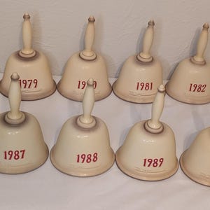 May include: A set of twelve cream-coloured ceramic bells with wooden handles. Each bell is inscribed with a year in red, ranging from 1978 to 1992. The bells are arranged on a white surface, showcasing their vintage design.