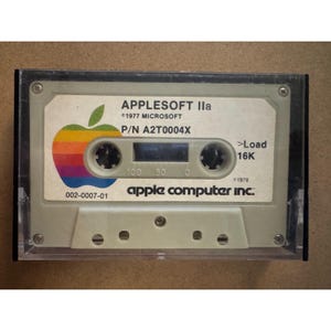 May include: A vintage beige cassette tape in a clear plastic case. The label features a rainbow-colored apple logo, the text APPLESOFT IIa, 1977 MICROSOFT, P/N A2T0004X, >Load 16K, 1978, 002-0007-01, and the apple computer inc. logo in black text.