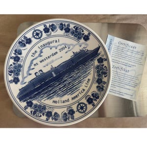 May include: A round white ceramic plate with blue floral patterns along the rim and a central illustration of the ms Westerdam cruise ship. The text reads the inaugural ms westerdam 2004 and Holland America Line. Includes a paper certificate.