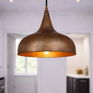May include: A copper-colored pendant light with a bell-shaped shade hangs above a marble countertop. The light fixture has a textured surface and a warm, golden interior. A bowl of lemons sits on the counter below.