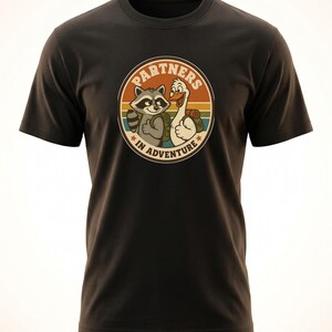 May include: Black t-shirt with a circular graphic showing a raccoon and a goose, both with backpacks, and the text "PARTNERS IN ADVENTURE". The design has a retro, outdoorsy aesthetic.