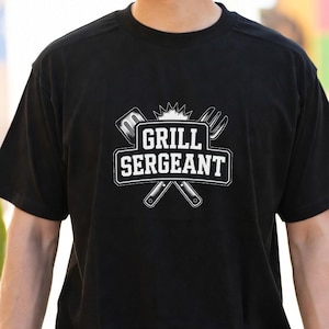Puede incluir: Camiseta negra con la inscripción blanca "GRILL SERGEANT". El diseño incluye una espátula y un tenedor cruzados detrás del texto, con una corona encima. La camiseta es de cuello redondo.