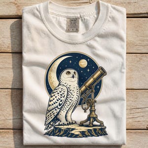 Può includere: Una t-shirt color crema con una stampa grafica di un gufo bianco in piedi su una sporgenza rocciosa accanto a un telescopio in ottone. Lo sfondo mostra un cielo notturno circolare blu scuro con una luna crescente, un pianeta e delle stelle.